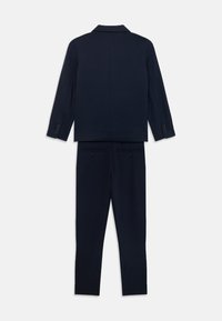 Mornarsko obleko, ki vključuje krojeno blazer s spodnjim ovratnikom in usklajenimi ožjimi hlačami, oboje z zadnjimi žepi in gumbnimi poudarki.