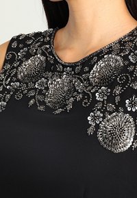 Černý oděv s jemným stříbrným květinovým vyš�íváním a ozdobami na kulatém výstřihu, ukazující texturu a podrobný design.