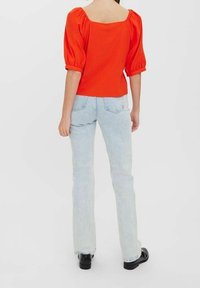 Haut rouge à manches bouffantes avec un décolleté carré, associé à un jean évasé délavé. Des chaussures noires complètent la tenue, sur un fond uni.