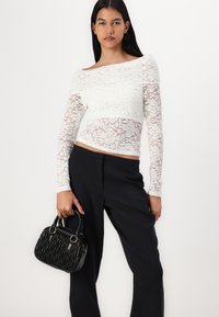 Haut en dentelle blanche à manches longues avec un design décolleté, associé à un pantalon noir ajusté. Tient un petit sac à main noir texturé.