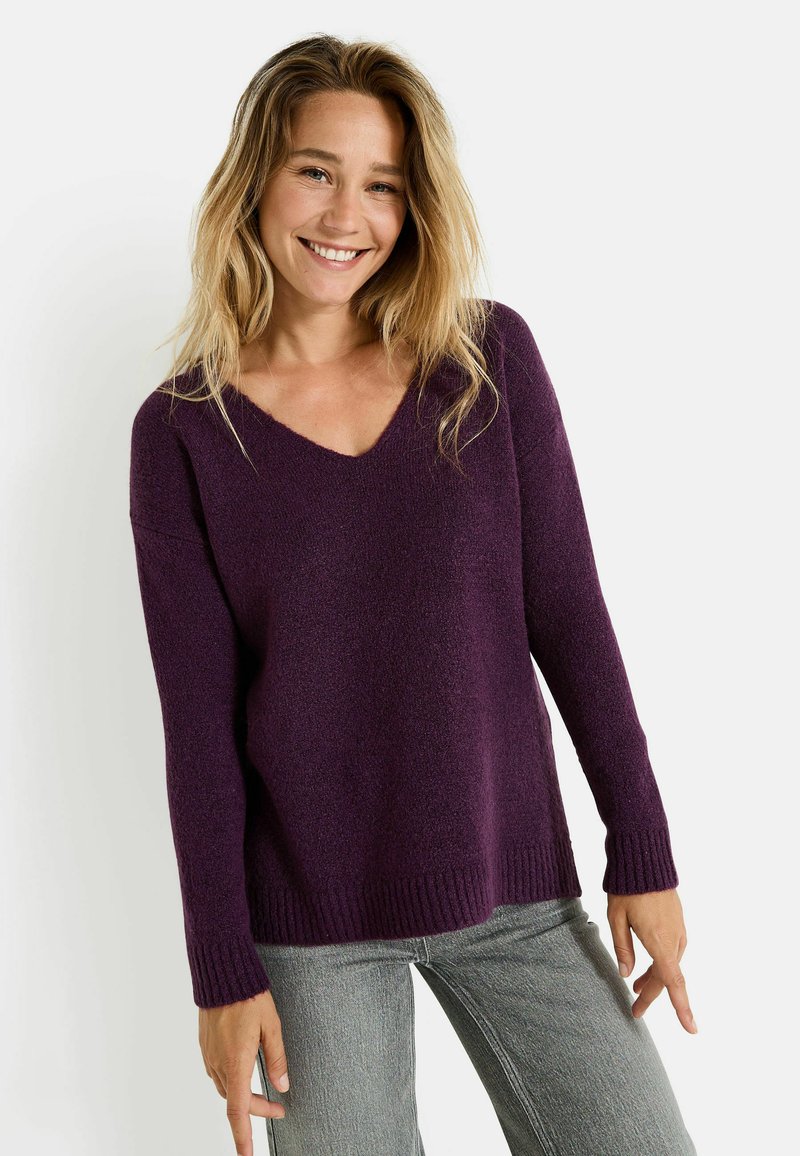 Suéter morado de cuello en V con un acabado texturizado, mangas largas y puños acanalados. La modelo lleva jeans grises y posa con las manos relajadas.