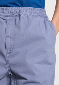 Närbild av en person som bär ljuslila byxor med elastisk midja, med en hand delvis i fickan, en blå skjorta synlig ovanför midjebandet.