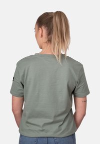 Camiseta de algodón color verde oliva con cuello redondo y mangas cortas; muestra de un pequeño logotipo negro en la manga izquierda; corte relajado.