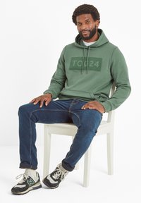 Olive green hoodie with a large TOC24 logo, featuring a drawstring hood, kangaroo pocket, and ribbed cuffs, paired with blue denim jeans and sneakers.