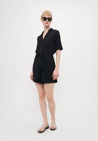 Svart kortermet jumpsuit med knapper foran, belte i midjen og to brystlommer. Modellen har på seg mørke solbriller og minimalistiske sandaler.