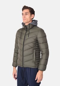 Giacca a vento verde oliva con zip frontale, motivo a zigzag e cappuccio grigio. Presenta polsini elasticizzati e tasche laterali. Tessuto liscio e isolante.