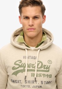 Felpa beige con cappuccio e coulisse, con logo e scritta "SuperDry" verdi. Indossata sopra una camicia a quadri verdi.