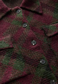 Wool jacket with a burgundy and green plaid pattern, featuring four black buttons and a collar. Textured fabric with a soft finish.
