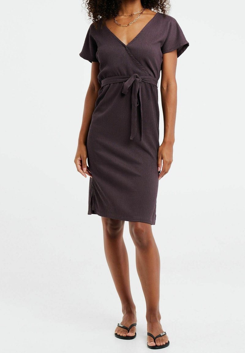 Femme portant une robe portefeuille marron foncé longueur genou avec manches courtes, une ceinture à la taille, un collier chaîne en or et des sandales tongs noires.