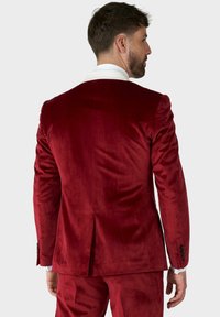 Roter Samtanzug mit einem einzigen Schlitz hinten, kontrastierendem weißen Kragen und schwarzen Knopfakzenten an den Ärmeln. Glatte Textur und maßgeschneiderte Passform.