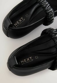 Zwarte instappers met een gladde leren afwerking, voorzien van een geplooide elastische band over de bovenkant en "NEXT MotionFlex" branding aan de binnenkant.
