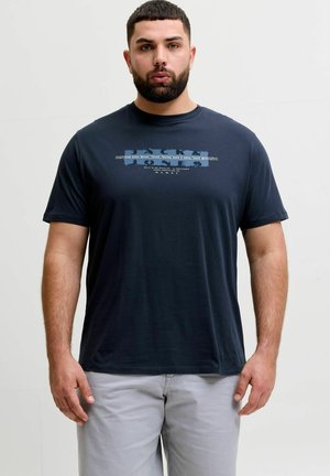 Homme barbu aux cheveux courts portant un t-shirt graphique bleu marine et un pantalon gris clair, debout devant un fond uni.