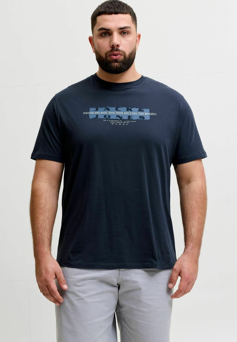 Homme barbu aux cheveux courts portant un t-shirt graphique bleu marine et un pantalon gris clair, debout devant un fond uni.