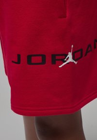 Shorts en coton rouge avec un logo "JORDAN" noir et un graphique silhouette Air Jordan blanc sur le côté inférieur gauche.