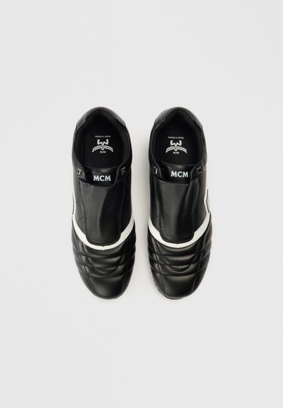 Zapatillas negras de cuero con un patrón acolchado, una franja blanca de acento, diseño sin cordones y el logo de MCM en la lengüeta y el lateral. Textura lisa.