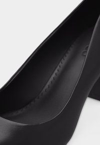 Chaussure noire à bout fermé à talon haut avec un talon bloc, présentant une finition mate lisse et un détail de couture intérieure.