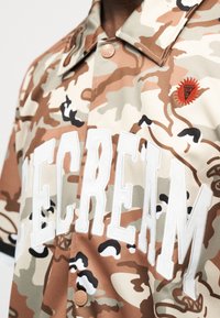 Μπλούζα παραλλαγής με έντονα λευκά γράμματα "ICECREAM" και μικρό κόκκινο έμβλημα ήλιου στο στήθος, με ορατό γιακά με κουμπιά.