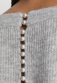 Grauer gestrickter Pullover mit einer Knopfverschluss auf der Rückseite, der aus sieben weißen, perlenähnlichen Knöpfen besteht, und einem strukturierten, weichen Stoff.