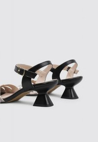Sandalias de tacón de cuero negro con recortes redondeados, una correa con hebilla y un distintivo tacón cónico acampanado. Textura suave y diseño minimalista.