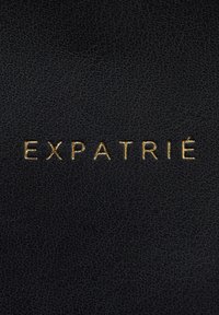 Černý kožený povrch s texturovaným finišem a razeným zlatým písmem, které píše "EXPATRIÉ" v moderním písmu.