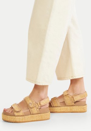 Beige Plateausandalen mit gewebter Strohtstruktur, verstellbaren Riemen und dicken Sohlen. Getragen mit cremefarbenen, weit geschnittenen Hosen.