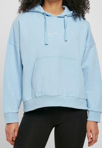 Ljust blå huvtröja av mjukt material, med en känguruficka, ribbade muddarna och en diskret broderad design på bröstet.