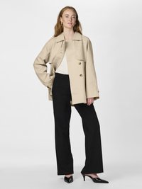 Beige overdimensjonert trenchcoat med store frontlommer og to lysebrune knapper, parret med svarte vide bukser og svarte spisse hæler.