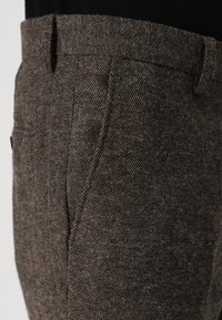 Rudos tweed kelnės su žuvelės kaulų raštu, turinčios tiesią kojų liniją ir tradicinį diržą. Tekstūruota medžiaga su subtiliomis pilkomis akcentais.