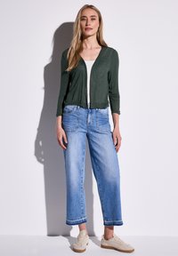 Grön lätt långärmad cardigan över en vit topp, i kombination med ljusblå vida jeans och beige sneakers, stående mot en vit bakgrund.