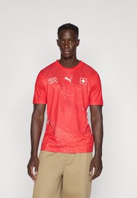 Piros sportos póló légáteresztő anyagból, texturált mintázattal, Puma logóval és svájci zászló jelvénnyel. Rövid ujjú, kerek nyakú.