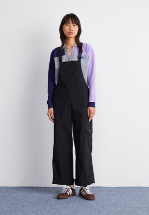 Black overalls with a loose fit, adjustable straps, and side pockets. Paired with a purple and grey long-sleeve top and brown sneakers.