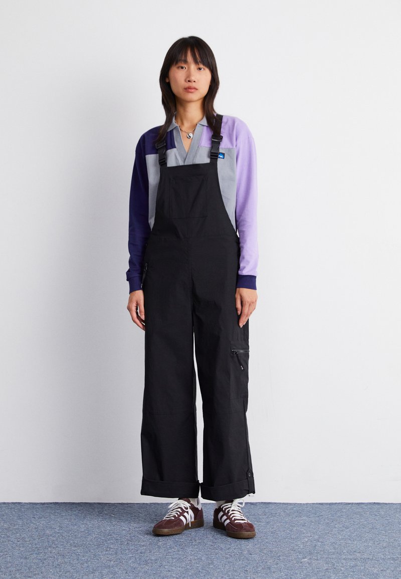 Black overalls with a loose fit, adjustable straps, and side pockets. Paired with a purple and grey long-sleeve top and brown sneakers.