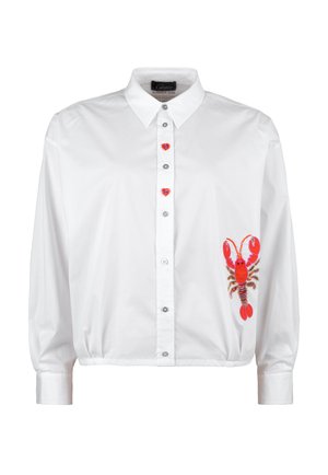 Smiling woman stands with hands in pockets wearing white shirt with red lobster graphic, light blue wide-leg jeans, and white sneakers.