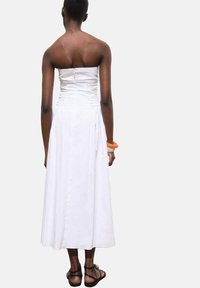 Witte strapless jurk met geplooide details op de buste, uitlopend in een knie-lange rok, gedragen met een eenvoudige oranje armband.