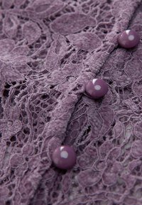 Lavendelspitzenstoff mit floralem Muster, der entlang des Randes drei glänzende, runde Knöpfe in derselben Farbe aufweist.