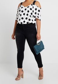 Donna che indossa jeans skinny neri a vita alta, top bianco con spalla scoperta a pois, sandali con tacco nudi, mentre tiene una pochette turchese con manico in metallo.