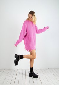 Överdimensionerad rosa stickad tröja med ribbat mönster och vågig nederkant. Bärs med svarta platåstövlar och vadlånga strumpor, mot en vit bakgrund.