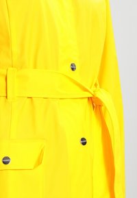 Gul regnrock i vattentätt material med bälte i midjan, tryckknappsdetaile och en framficka. Slät textur med en ljus finish.