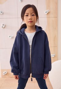 Sweat à capuche zippé bleu marine avec capuche, poches avant et poignets élastiques, associé à un t-shirt blanc et un pantalon de sport doux.