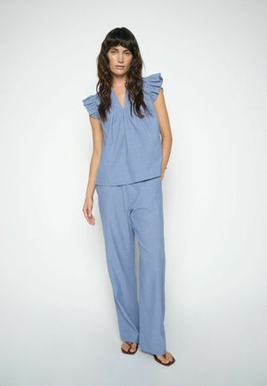 Hosszú sötét hajú nő világoskék ujjatlan blúzt visel fodros vállrésszel és hozzá illő, bő szárú nadrágot, egyik kezét a zsebében tartva áll.