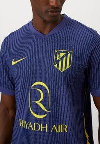 Marineblauwe voetbalshirt met textuurdesign, V-hals, gele Nike-logo, clubembleem, en "RIYADH AIR" in het geel gedrukt.