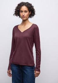 Burgunderfarbenes Langarm-T-Shirt mit V-Ausschnitt und weicher, glatter Textur. Kombiniert mit blauen Jeans, die einen lässigen, entspannten Schnitt haben.