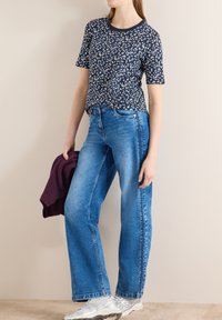 Femme portant une chemise à manches courtes bleu marine à motifs, un jean large bleu et des baskets argentées, tenant un pull violet plié dans la main gauche.