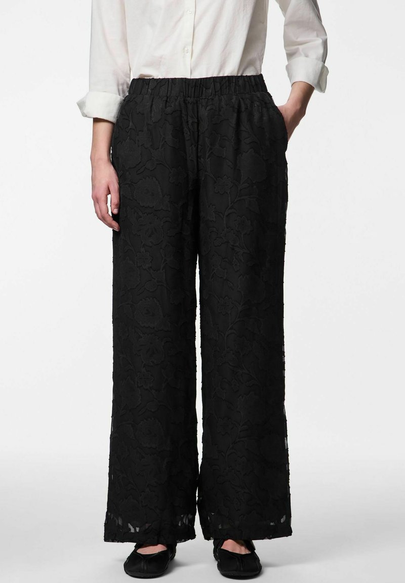 Mustad tikanditega laiade säärtega püksid, millel on elastne vöökoht, lillemustrid ja läbipaistev pealmine kiht. Kandes neid koos mustade madalate kingadega.