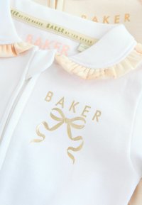 Balta bērnu krekla ar ruflētu persiku krāsas apkakli, uz kuras redzams zelta BAKER logo un loku dizains. Mīksta kokvilnas auduma, gluda tekstūra.