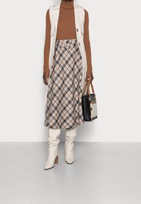 Beige ärmellose Weste über einem braunen gerippten Rollkragen, kombiniert mit einem karierter Rock in braunen und weißen Tönen. Weiße Schlabberstiefel, schwarze Handtasche.