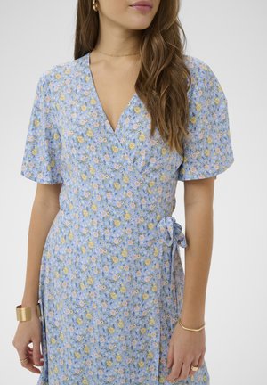 Femeie purtând o rochie înfășurată cu flori bleu deschis, cu mâneci scurte, cercei rotunzi aurii, brățară și inele, părul lăsat liber peste un umăr.