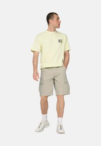 Camiseta corta de manga amarilla claro con diseño de bolsillo a cuadros, combinada con pantalones cortos de cargo beige y zapatillas blancas.