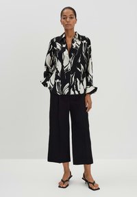 Camicia a righe bianco e nero con un design astratto fluente, abbinata a pantaloni larghi neri e sandali infradito neri.