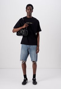 Camiseta negra de manga corta con texto dorado, combinada con pantalones cortos de denim azul desgastados. El modelo sostiene un bolso negro brillante. Zapatillas negras y calcetines.